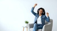 Happy young African American woman dancing with headphones in modern chair celebrating music and joy at home.