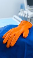 Orange latex glove resting on blue medical drape in clinical setting with ultrasound equipment blurred in background, representing healthcare, safety, and medical examination procedures.
