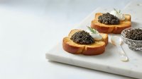 Toasted bread slices topped with creamy spread and black caviar garnished with fresh herbs on white marble board, gourmet appetizer.