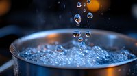 Macro photography of water drops falling into glass with crystal clear liquid, creating splash crown formation against dark background with orange bokeh lights.