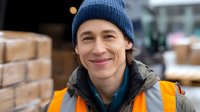 Young warehouse worker in blue beanie and safety vest smiling outdoors near loading dock area, expressing positive attitude during winter workday.