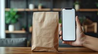 Hand holding modern smartphone with blank screen next to kraft paper bag on wooden table, mockup for delivery app or food ordering service.