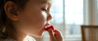 Young girl with medium skin tone eating fresh red strawberry by window with natural daylight, suitable for food, health, nutrition, and lifestyle content.