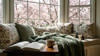 Cozy reading nook with open book and coffee mug by window overlooking blooming cherry blossom trees in spring creating peaceful home interior atmosphere.
