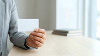 Young adult male with medium skin tone holding blank white business card at wooden desk, wearing casual gray sweater, professional networking and corporate identity concept.