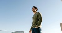 Young Asian male wearing olive green hoodie and dark jeans standing outdoors against clear sky, looking upward with confident expression, suitable for lifestyle and casual apparel marketing.