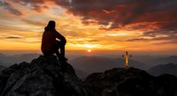 Person silhouette sitting on mountain peak at sunrise with cross on distant hill, spiritual contemplation and faith concept for religious meditation designs.