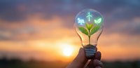 Green energy lightbulb with plant and wind turbine inside held against sunset sky representing sustainable innovation and renewable power solutions.
