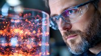 Bearded Caucasian man in glasses observing glowing particles in glass container, side view portrait against dark background with bokeh lights and scientific experiment.