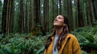 Young woman in mustard cardigan smiling while looking up in lush green forest with tall trees and ferns, conveying joy and connection with nature.