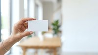 Hand holding blank white card in modern bright interior with wooden furniture and green plant, suitable for business branding and marketing mockups.