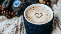 Cozy coffee cup with heart-shaped foam art in dark blue knitted cup holder surrounded by scattered coffee beans on textured fabric background.