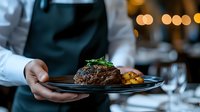Professional chef presenting grilled beef steak with herbs and roasted potatoes on black plate in upscale restaurant kitchen with warm ambient lighting.