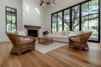 Modern bright living room with rattan chairs, white sofa, hardwood floors and large windows overlooking garden. Natural light streams through black-framed windows. Minimalist design.