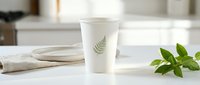 Disposable white paper cup with fern leaf design on modern kitchen counter with fresh green leaves and ceramic plate for beverage branding mockup.