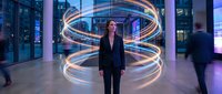 Woman in business suit standing in modern office lobby with glowing neon spiral light trails, surrounded by glass buildings and blurred figures, representing innovation and corporate technology.