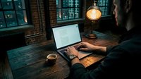Young Caucasian man working on laptop at rustic wooden desk with glowing lamp and coffee cup in moody urban loft office at night.
