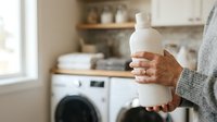 Mature adult with medium skin tone holding white laundry detergent bottle in modern kitchen with washing machine, representing household cleaning and laundry care routines.