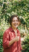Happy young Caucasian woman giving thumbs up gesture in red shirt outdoors among green foliage with natural sunlight creating cheerful positive mood.
