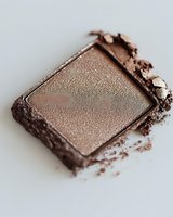 Crushed metallic brown eyeshadow palette with scattered powder showing texture and shimmer detail on white background, macro cosmetic photography.