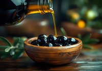 Pouring fresh olive oil over black olives in wooden bowl on rustic table with green leaves. Dark moody food photography with natural lighting.