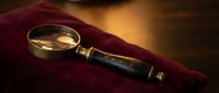 Vintage magnifying glass with brass lens and dark leather-wrapped handle resting on deep burgundy fabric, warm golden lighting for antique collecting, detective work, or mystery themes.