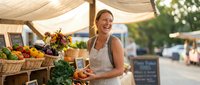 Young Caucasian woman vendor laughing while holding fresh tomatoes at outdoor farmers market stall with organic vegetables and fruits under canvas tent.