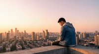 Young man in denim jacket sitting on rooftop edge overlooking urban skyline at sunset, contemplating city view from high building terrace during golden hour.