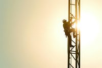 Silhouette of telecommunications tower climber working at height during sunset, industrial worker maintaining cellular antenna equipment with safety harness.