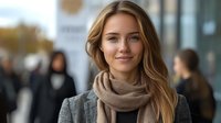 Young blonde businesswoman wearing beige scarf and gray coat smiling confidently on urban street with blurred people walking in background during autumn day.