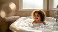 Young mixed-race girl with curly hair enjoying bubble bath in modern bathroom with warm sunlight streaming through window creating cozy atmosphere.