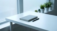 Minimalist white desk with closed laptop and pen near window with potted plants creating clean modern workspace for productivity and focus.