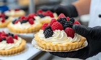 Fresh berry tartlets with whipped cream, raspberries and blackberries on silver doily, held by hand in black glove. Pastry chef preparing desserts in bakery.