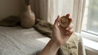 Mature hand holding vintage brass compass near window with natural light, linen curtains, and ceramic vase for travel, navigation, or lifestyle concepts.
