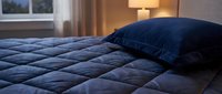 Cozy bedroom interior with navy blue quilted comforter and matching pillow on bed, warm lamp lighting, window with curtains, suitable for home decor and hospitality design projects.