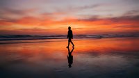 Silhouetted male surfer walking along wet beach sand at golden hour sunset, carrying surfboard under arm, reflected in water, coastal seascape.