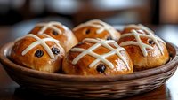 Fresh hot cross buns with raisins in rustic woven basket, traditional Easter bread rolls with glazed cross pattern on top, close up view of baked pastries.