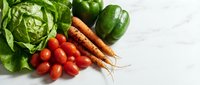 Fresh organic vegetables including leafy greens, cherry tomatoes, carrots and bell pepper arranged on white marble surface for healthy cooking and meal prep.