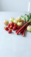 Fresh organic produce arrangement with green apples, red strawberries and rhubarb stalks on clean white surface for healthy cooking and nutrition concepts.