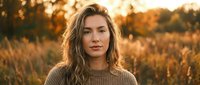 Young blonde woman in cozy sweater standing in golden autumn field during sunset with warm bokeh lights creating dreamy atmospheric portrait for lifestyle content.