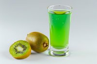 Bright green kiwi juice in tall shot glass with fresh whole and sliced kiwi fruit on light background, refreshing summer drink.
