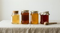 Four glass jars filled with different varieties of honey in golden and amber tones arranged on linen tablecloth with wooden dipper for organic food concepts.