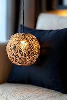 Modern spherical woven rattan pendant lamp glowing warmly against dark blue pillow background, creating cozy atmosphere in contemporary living space.