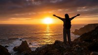Silhouetted figure with arms raised celebrating success on rocky coastal cliff at golden sunset over ocean waters.
