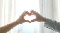 Two people making a heart shape with their hands against a bright window, symbolizing love, unity, and togetherness in a warm, soft light.