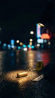 Bullet casing on wet asphalt at night with evidence marker number three, bokeh city lights in background, crime scene investigation.