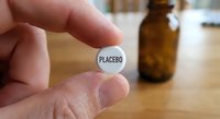Hand holding white placebo pill with amber medicine bottle in background on wooden surface for pharmaceutical research and clinical trial concepts.
