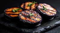 Grilled eggplant slices with rosemary herbs and balsamic glaze on black stone background, close up view of roasted vegetables with charred marks.