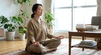 Young Asian woman practicing meditation in lotus position at home with houseplants creating peaceful wellness atmosphere for mindfulness and relaxation.