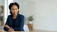 Confident African American female professional in navy blazer seated at desk with arms crossed, displaying leadership and corporate authority in modern office environment.